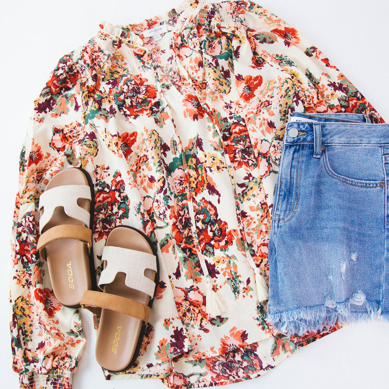 Cream blouse with multi color floral print featured with denim shorts and neutral sandals on a white backdrop