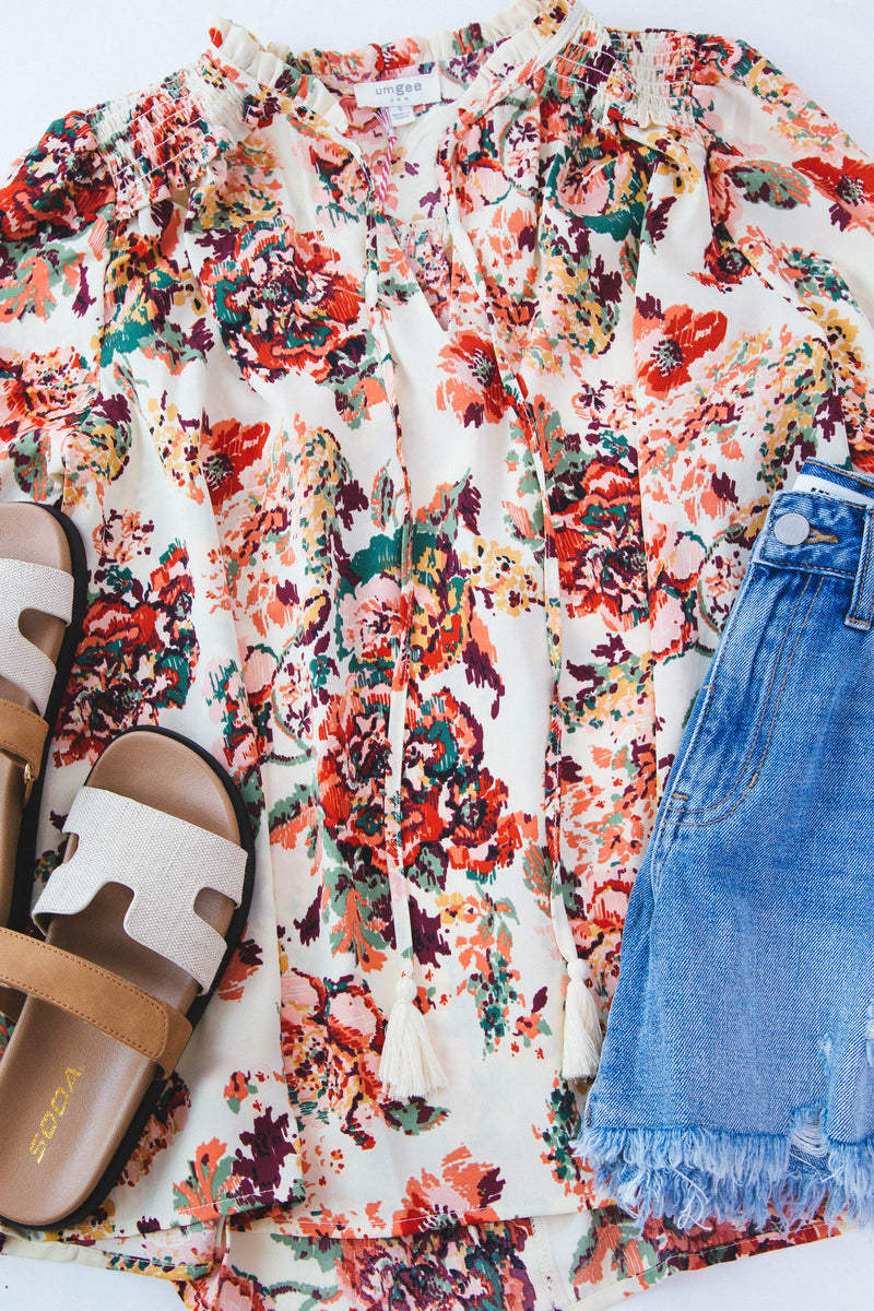 Cream blouse with multi color floral print featured with denim shorts and neutral sandals on a white backdrop