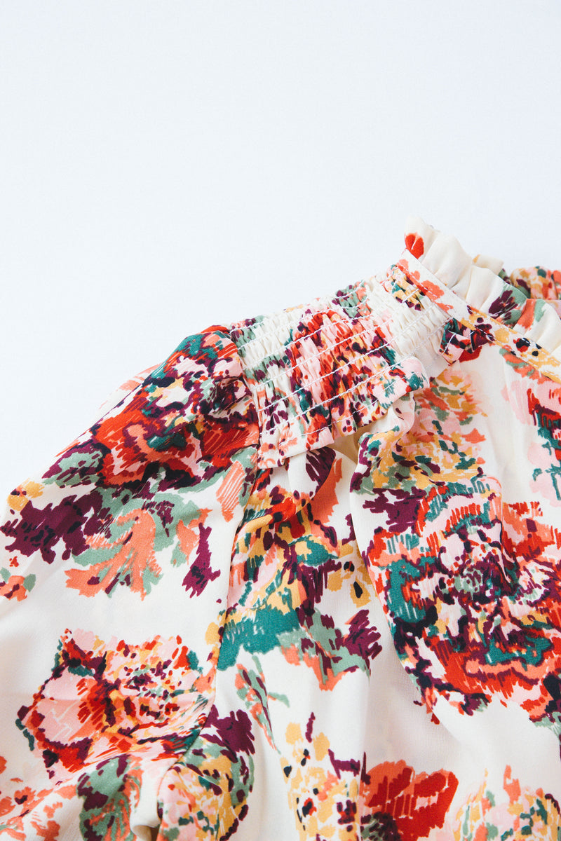 Cream blouse with multi color floral print featured with denim shorts and neutral sandals on a white backdrop ; detail shot